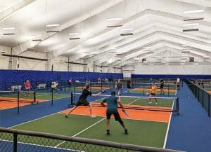 Group of people at a Dill Dinkers indoor pickleball grand opening ceremony, standing in front of a branded backdrop featuring the Dill Dinkers and JOOLA logos. They are holding a blue ribbon labeled 'GRAND OPENING,' marking the launch of a new pickleball facility.