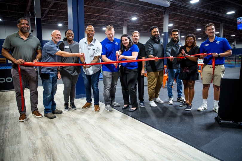 Group of people holding a red ribbon in an indoor Dill Dinkers pickleball facility, preparing for a ribbon-cutting ceremony. The space features high ceilings and overhead lighting, marking the launch of a new indoor pickleball location.