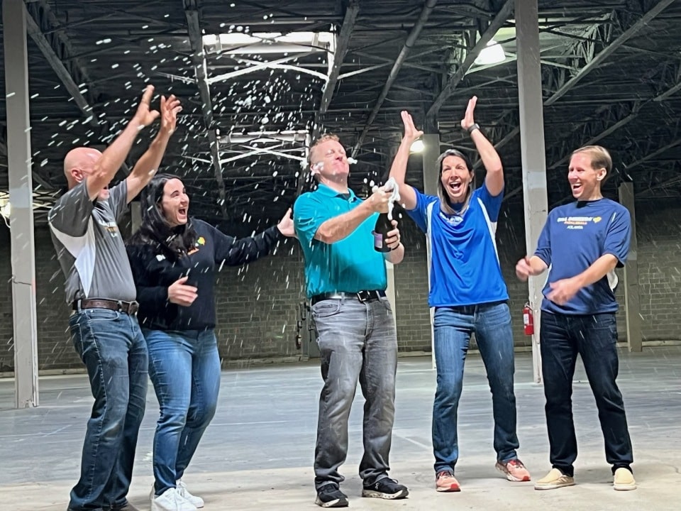 Celebration at a Dill Dinkers indoor pickleball facility with five people in casual clothing standing in an industrial-style space. One person sprays champagne into the air under high ceilings with exposed beams, marking a milestone for the Dill Dinkers pickleball brand