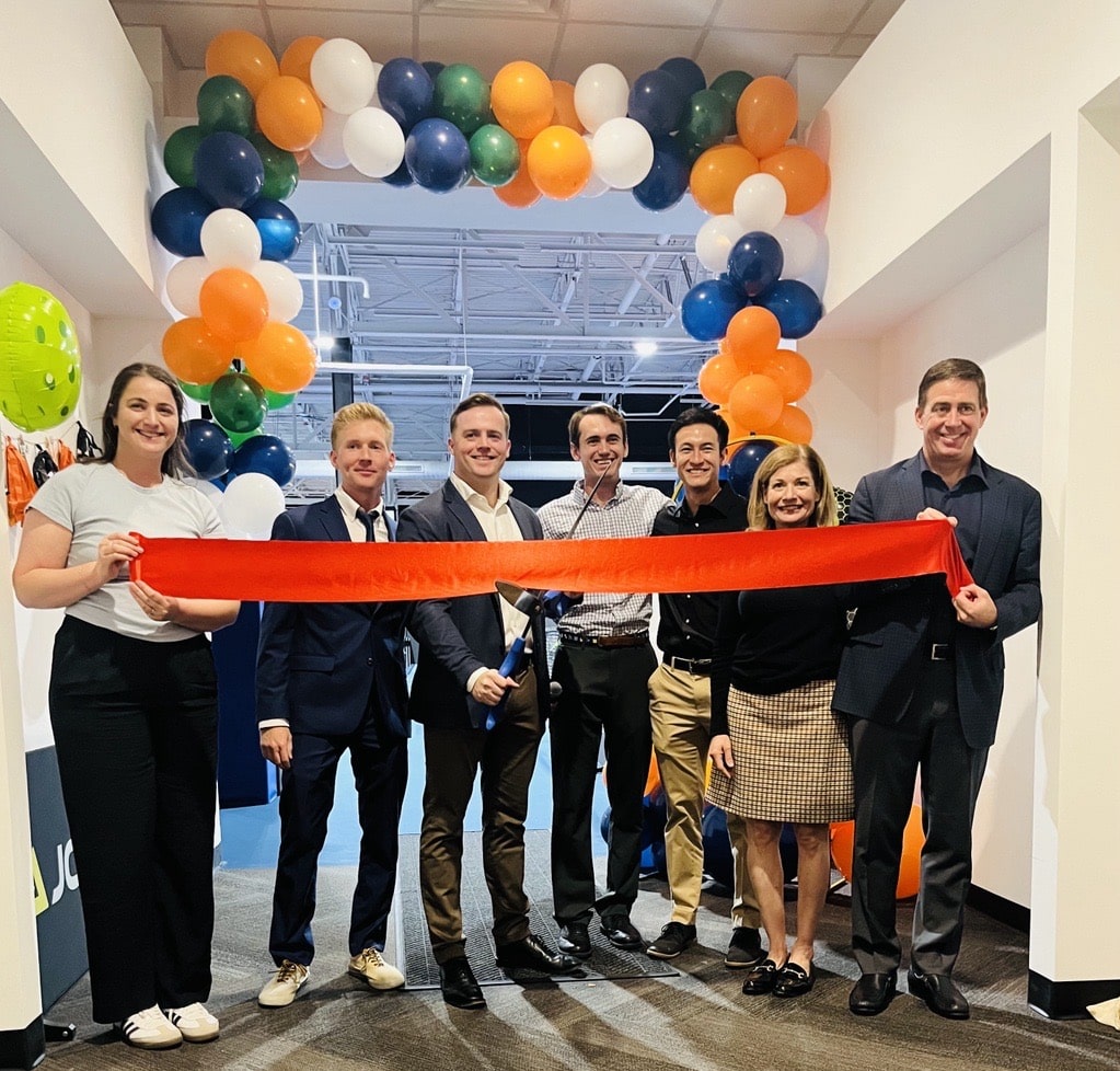 Group of people at a ribbon-cutting ceremony standing under a colorful balloon arch made of orange, green, blue, and white balloons. The event appears to mark the opening of a new Dill Dinkers facility.