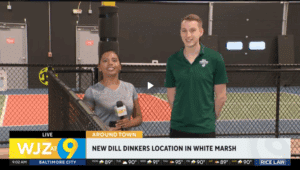 A local news segment shows two people standing inside an indoor pickleball facility. One person is holding a microphone with a WJZ logo, and the other is wearing a green polo shirt with a Dill Dinkers logo. Behind them are pickleball courts enclosed by black fencing. The on-screen text reads “LIVE WJZ at 9 Baltimore City,” “Around Town,” and “New Dill Dinkers location in White Marsh.” The bottom bar also displays the time, weather forecast, and sponsor information.