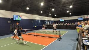 Four players engaged in a doubles pickleball match on an indoor court, with two players near the net and two positioned farther back. Spectators are seated along the side behind a black barrier, and scoreboards are visible on the dark blue walls.